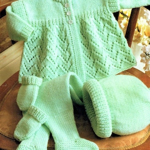 Könnte beinhalten: Ein grünes gestricktes Baby-Outfit mit einem Cardigan, Leggings und einer Mütze. Der Cardigan hat einen Knopfverschluss und ein Spitzenmuster.