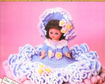 May include: A crocheted doll wearing a blue dress with white and pink ruffles, a blue hat with white ruffles and pink flowers. The doll has brown hair and blue eyes. The doll is sitting on a pink and white crocheted base. Free Gift Tags.