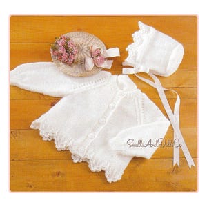 May include: A white knitted baby outfit set, including a cardigan, bonnet, and a small hat with pink flowers and a ribbon. The cardigan has button closures and a scalloped edge. The text "Small's And Dolls Co" is visible.