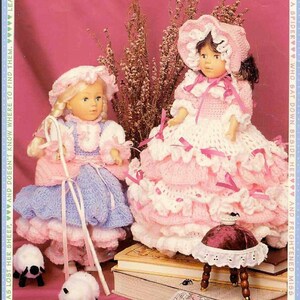 May include: Two crocheted dolls dressed in pink and blue dresses with white lace trim. The dolls are holding a shepherd's crook and a small stool. The dolls are sitting on a stack of books with a small white sheep in front of them.