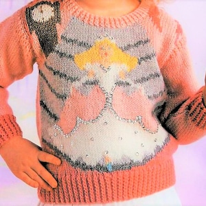May include: A pink knitted jumper with a white and grey design of a fairy with blonde hair and a sparkly dress. The jumper has a crew neck and long sleeves.