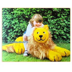 May include: A young girl sits on a large, yellow plush lion. The lion has a friendly face with black eyes and a black nose. The girl is wearing a white shirt and blue jeans. The lion and girl are sitting on a green grassy lawn.