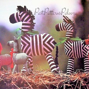May include: Four stuffed zebra toys, two large and two small, with black and white stripes. The larger zebras have green ribbon accents. The smaller zebras are red and green. The zebras are sitting on a bed of straw.