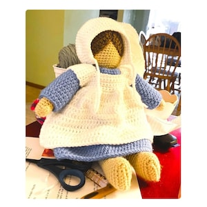 May include: A crocheted doll wearing a white dress and blue bonnet. The doll has brown yarn hair and is sitting on a table with a pair of scissors and a notebook.