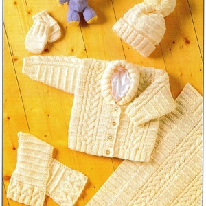 Op de afbeelding: Een gebreide babyset in crème met een kabelbreipatroon. De set bevat een trui, muts, slofjes en een deken.