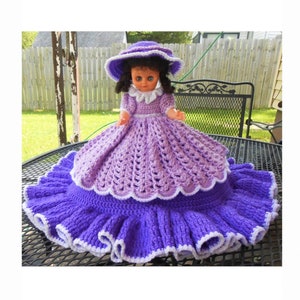 Puede incluir: Un vestido de muñeca de ganchillo morado con ribete blanco. El vestido tiene una falda con volantes y un corpiño ajustado. La muñeca lleva un sombrero morado con ribete blanco.