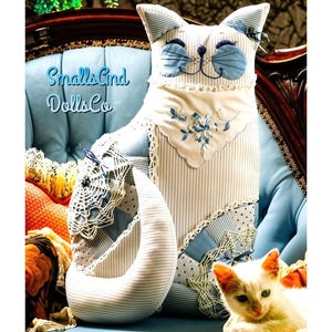May include: A fabric cat-shaped pillow featuring a light blue and white pinstripe pattern, accented with a white lace collar, embroidered floral details, and a crocheted doily on the tail. The text Smalls And DollsCo is visible on the left side.