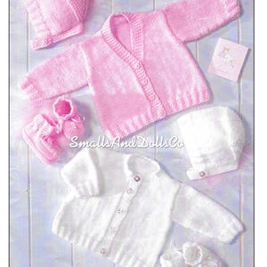 Op de afbeelding: Twee gebreide baby-outfits, een roze en een witte. Elke outfit bevat een trui, een muts en slofjes. De roze outfit heeft een witte knoopsluiting en de witte outfit heeft een zilveren knoopsluiting.