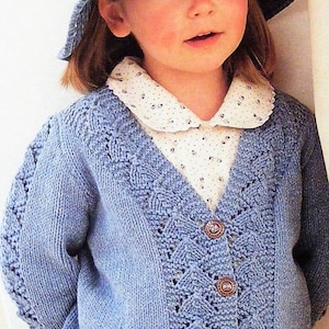 Op de afbeelding: Een jong kind draagt een lichtblauwe gebreide cardigan met een wit overhemd met kraag eronder. De cardigan heeft een kantpatroon en wordt vastgemaakt met bruine knopen. Het kind draagt ook een denimhoed.