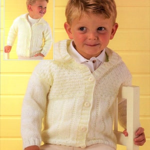 May include: A white knitted cardigan with a button fastening and a collar. The cardigan is being worn by a young boy.