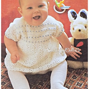 May include: A smiling baby wearing a white crocheted dress with blue accents and white tights. A stuffed bunny and a colorful toy are in the background. The text "Small Cind Dolls Co" is visible.