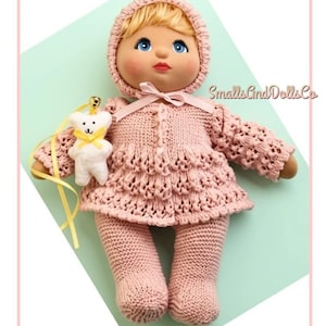 Può includere: Una bambola con capelli biondi e occhi azzurri indossa un cappellino di lana rosa e un vestito rosa testurizzato abbinato con orlo a balze. La bambola tiene in mano un piccolo orsetto di peluche bianco con un nastro giallo e un campanellino.