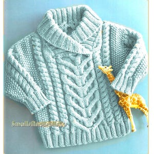 May include: A light blue cable-knit sweater with a wide, ribbed collar and sleeves. The sweater features a central cable design and the text "SmallsAndDollCo". A small giraffe figurine is placed on the sweater.