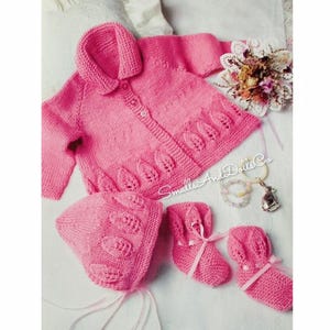 May include: A pink knitted baby set including a collared cardigan, bonnet, and booties. The set features leaf-shaped cutouts and is tied with pink ribbons. The cardigan has three buttons. The set is displayed on a white surface.
