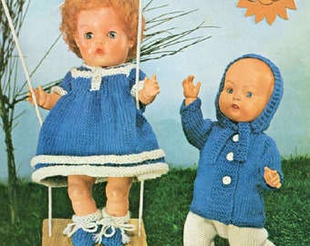 Op de afbeelding: Twee gebreide poppenoutfits in blauw en wit. De meisjespop draagt een blauwe jurk met witte bies en witte schoenen. De jongenspop draagt een blauwe trui met witte knopen en een witte broek met blauwe schoenen. Beide poppen hebben een vintage look.