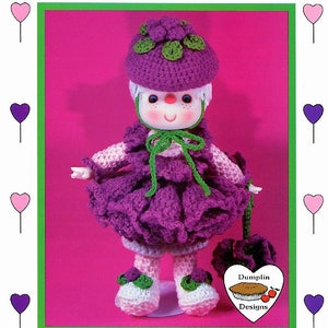 May include: Crocheted doll with a purple dress, green hat, and white shoes. The doll is holding a green and purple flower. The doll is labeled "Raspberry Fluff" and "Dumplin Designs".