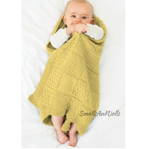 May include: A yellow knitted baby blanket with a square pattern. The blanket is wrapped around a baby lying on a white surface. The text "SmallAndDolls" is visible in the lower right corner of the image.