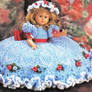May include: A crocheted doll dress in blue and white. The dress has a ruffled hem and is decorated with small, crocheted flowers. The doll is wearing a white hat with a red bow.