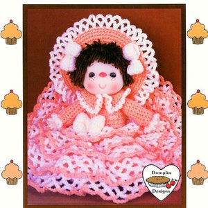 May include: A crochet doll wearing a pink and white dress and a white hat. The doll has brown hair, blue eyes, and a pink and white dress. The doll is sitting on a pink and white crocheted blanket. The doll is labeled "Peach Taffy" and "Dumplin Designs".