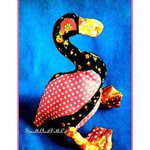 Può includere: Una bambola a forma di cigno in tessuto fatta a mano, con collo e testa a fantasia floreale nera, corpo a pois rosa e dettagli gialli e arancioni. Il testo "SmallsAndDollsCo" è visibile su sfondo blu.