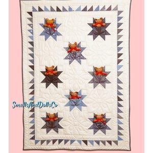 May include: A handmade quilt featuring a repeating star pattern with a teddy bear in each star. The quilt has a white background with blue and gray accents. The border is a triangle pattern. The text "SmallsAndDollsCo" is visible.
