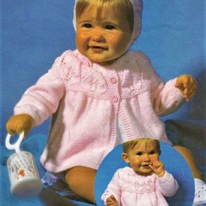 May include: A pink knitted baby cardigan with a button closure and a delicate lace pattern. The cardigan is worn by a baby wearing a pink knitted bonnet.