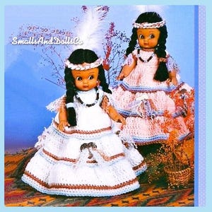 May include: Two crocheted dolls dressed in Native American inspired outfits. The doll on the left is wearing a white dress with brown and blue trim. The doll on the right is wearing a pink dress with blue trim. Both dolls have black hair and brown eyes.