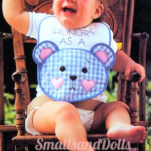 Könnte beinhalten: Ein Baby trägt eine blaue und weiße Gingham-Lätzchen mit einem rosa und blauen Bären-Design. Das Lätzchen hat den Text "Hungry as a..." gestickt. Das Baby sitzt in einem Holz-Schaukelstuhl.