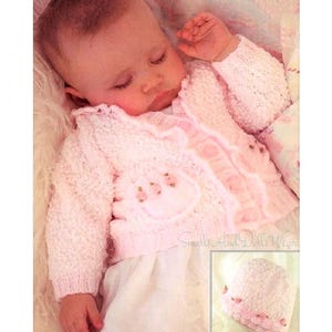 Könnte beinhalten: Ein rosa gestrickter Baby-Cardigan mit Rüschenbesatz und einem Blumendesign auf der Vorderseite. Der Cardigan ist auf einem schlafenden Baby auf einer weißen Decke abgebildet. Der Text "Smalls And Dolls Wear" ist in der rechten unteren Ecke des Bildes sichtbar.