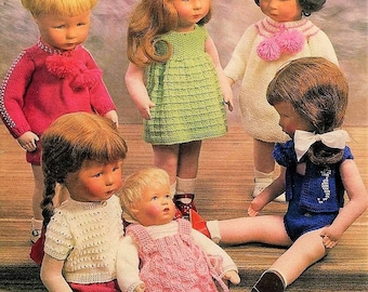 Op de afbeelding: Zes poppen die gebreide kleding dragen. De poppen zitten en staan op een houten oppervlak. De poppen dragen verschillende kleuren, waaronder roze, groen, blauw, wit en rood.