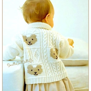 Puede incluir: Cárdigan de bebé color crema con tres caras de osito de peluche bordadas en la espalda. El cárdigan tiene un diseño de punto trenzado y un dobladillo acanalado. El bebé lleva una falda beige. El texto "Smalls And Dolls Co" es visible.