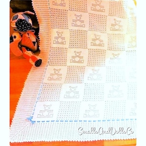 May include: White crocheted baby blanket with a blue border and a repeating pattern of small bears. The blanket is on a wooden surface with a stuffed animal toy in the corner.