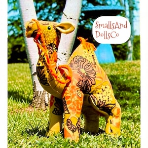May include: A stuffed camel toy made of yellow fabric with a brown and orange floral pattern. The camel features a bright orange yarn bridle and a small orange yarn tuft on its back. A white label with the text SmallsAnd DollsCo is visible.
