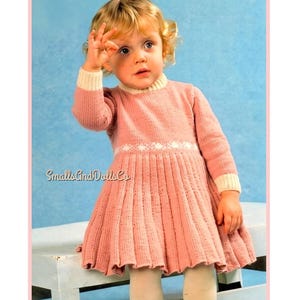 May include: A long-sleeved, dusty pink knitted dress featuring a ribbed texture, a cream-coloured collar, and cream-coloured cuffs. A horizontal white band with a delicate floral pattern encircles the waist. The text SmallsAndDollsCo is visible.