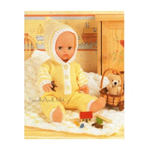 May include: A yellow knitted baby doll outfit with white stripes, a matching hat, and a button closure. The doll is sitting on a white blanket with a teddy bear in a basket and other toys.