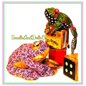 May include: Two fabric frog toys, one green with black polka dots and one purple with a floral pattern, sitting on top of wooden blocks. The blocks have a label that reads "Smoking Feat".