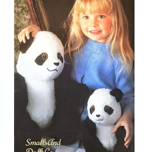 May include: Two plush panda toys with white and black fur, brown eyes, and stitched facial features. The larger panda is positioned on the left, and the smaller panda is on the right. The text SmallsAnd DollsCo is printed in gold on the lower left.