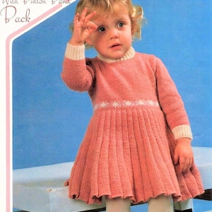 Puede incluir: Un vestido de punto rosa con ribete blanco y una banda de botones. El vestido es plisado y tiene mangas largas. El vestido lo lleva puesto un niño pequeño.