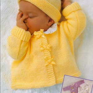 May include: A yellow knitted baby cardigan with a bow detail and matching hat. The cardigan has a button closure and is made with a soft yarn. "Smalls And Dolls Wear" is printed on the bottom of the image.