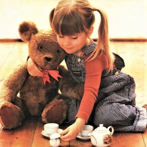 May include: A young girl in a red shirt and blue overalls sits on the floor with a brown teddy bear. She is holding a teacup and saucer in one hand and a teapot in the other. There are three teacups and saucers on the floor in front of her.