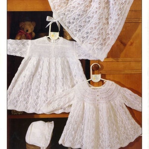 Op de afbeelding: Drie witte gehaakte babykledingstukken: een jurk, een vest en een muts. De kledingstukken worden getoond op een houten plank met een teddybeer op de achtergrond.