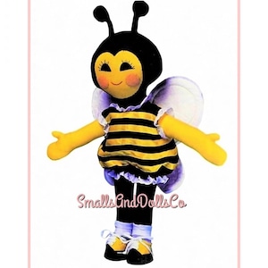 May include: A plush bee doll with a black and yellow striped shirt, black limbs, and white wings. The doll features a black head with antennae, pink cheeks, and long eyelashes. The text SmallsAndDollsCo is printed in pink across the lower torso area.