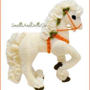 May include: A white crocheted horse toy with a textured mane and tail, featuring an orange harness with small yellow dots and green leaf accents. The horse has dark brown hooves and black eyes. The text SmallsAndDollsCo is visible on the left.