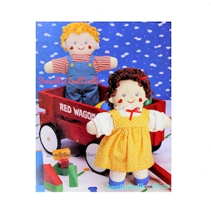 May include: Two handmade fabric dolls, a boy and a girl, are sitting in a red wagon with a white wheel. The boy doll is wearing a red and white striped shirt and blue overalls. The girl doll is wearing a white blouse with a yellow polka dot dress. The wagon has the words "Red Wagon" printed on the side.