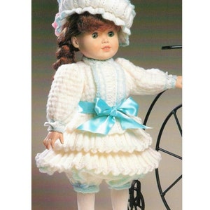Puede incluir: Un conjunto de muñeca de punto blanco con una cinta azul y un gorro de punto blanco. La muñeca lleva calcetines blancos y zapatos blancos con detalles azules.
