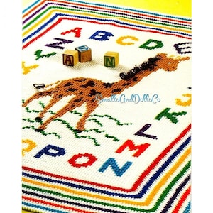 May include: A crocheted baby blanket with a giraffe design and the alphabet. The blanket features a white background with a colorful border and letters. Wooden blocks with letters are placed on the blanket. The text "Smalls And Dolls Co" is also visible.