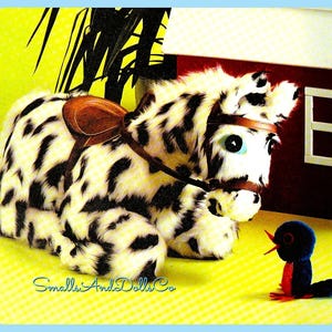 Op de afbeelding: Een zwart-wit gevlekt pluche schommelpaard met een bruin zadel en hoofdstel. Het paard ligt en kijkt naar een kleine blauwe vogel met een rode snavel en oranje poten.