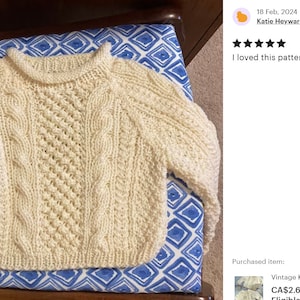 May include: A white knitted baby sweater with a cable knit pattern. The sweater is laying on a blue and white patterned surface.