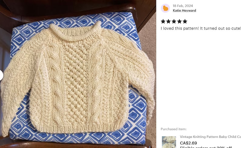 May include: A white knitted baby sweater with a cable knit pattern. The sweater is laying on a blue and white patterned surface.
