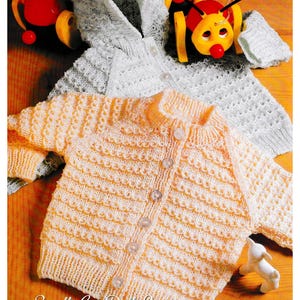 Könnte beinhalten: Zwei gestrickte Baby-Pullover, einer in hellgrau mit Kapuze und der andere in Pfirsichfarbe mit Knöpfen. Die Pullover liegen auf einer Holzoberfläche mit einem Spielzeug im Hintergrund.